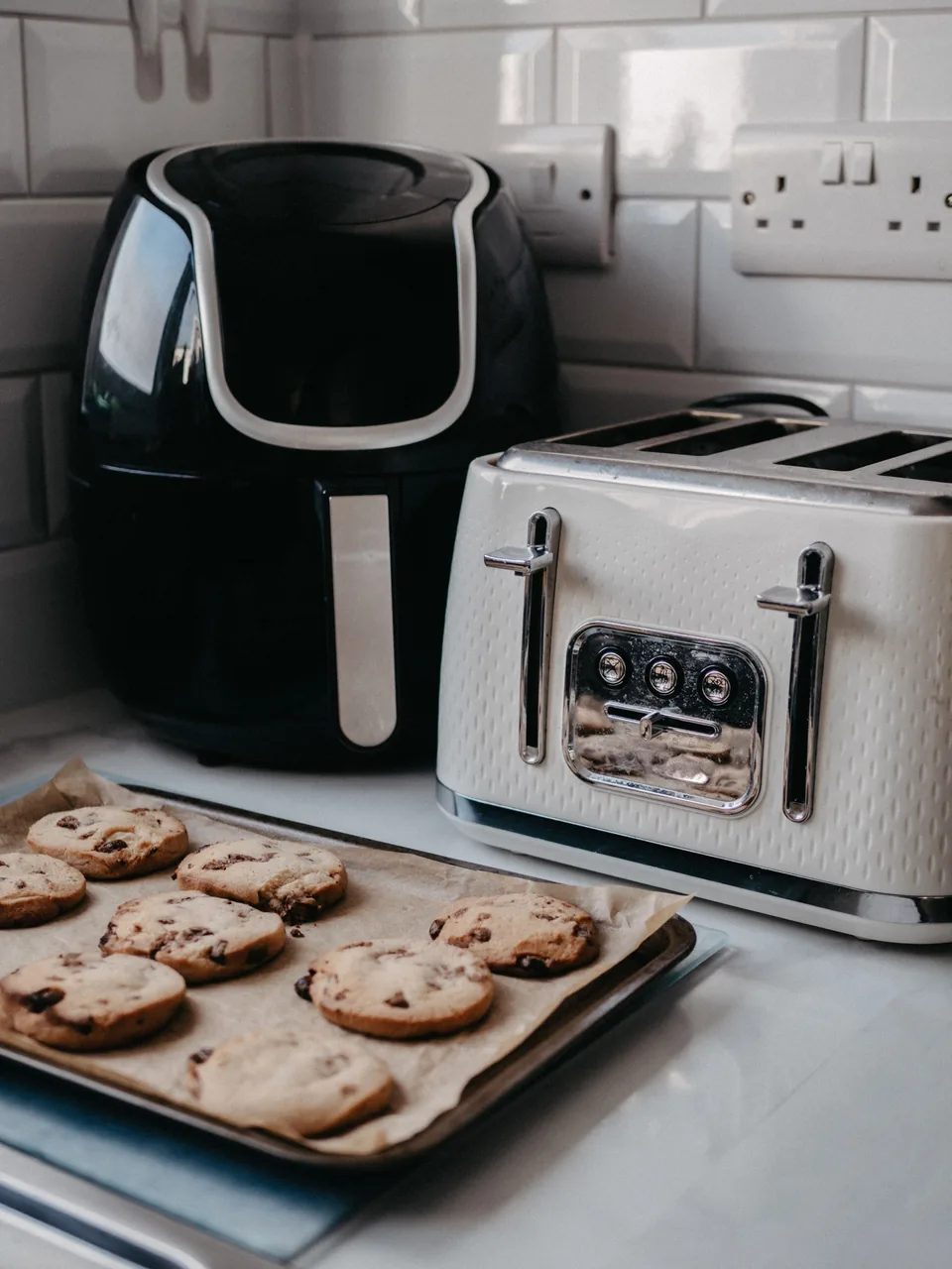 Black air fryer beside a toaster and a tray of cookies on a kitchen counter.